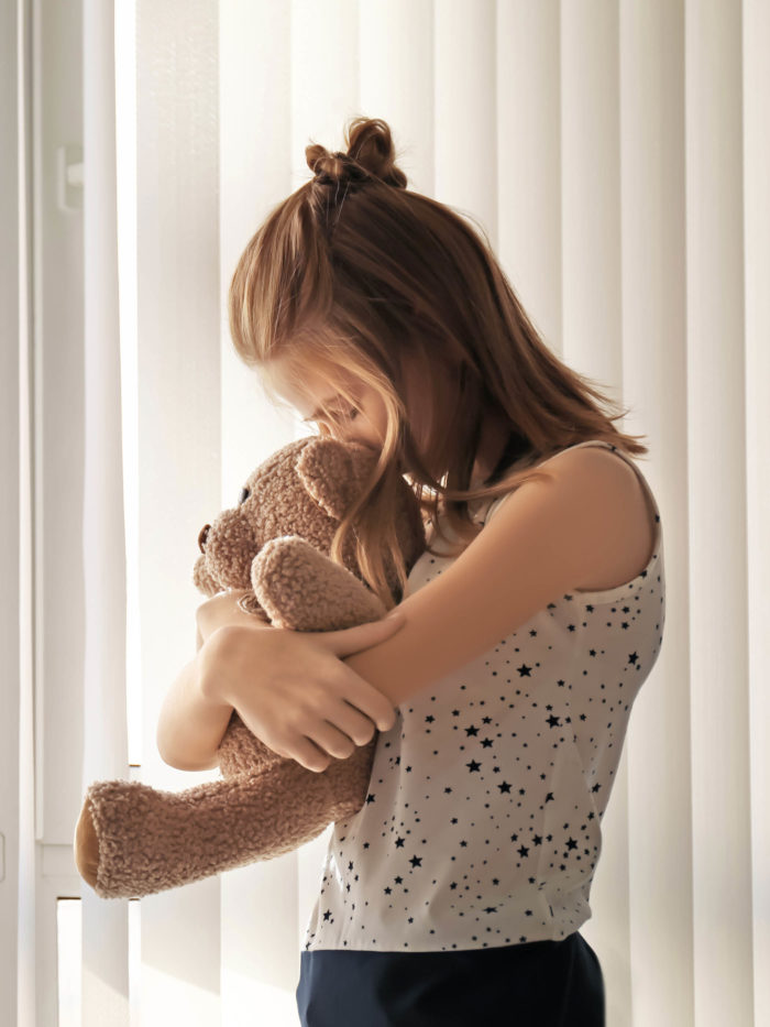 young girl with pensive look holding eddy bear near window