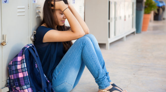 teen girl crying sitting on floor by lockers at school