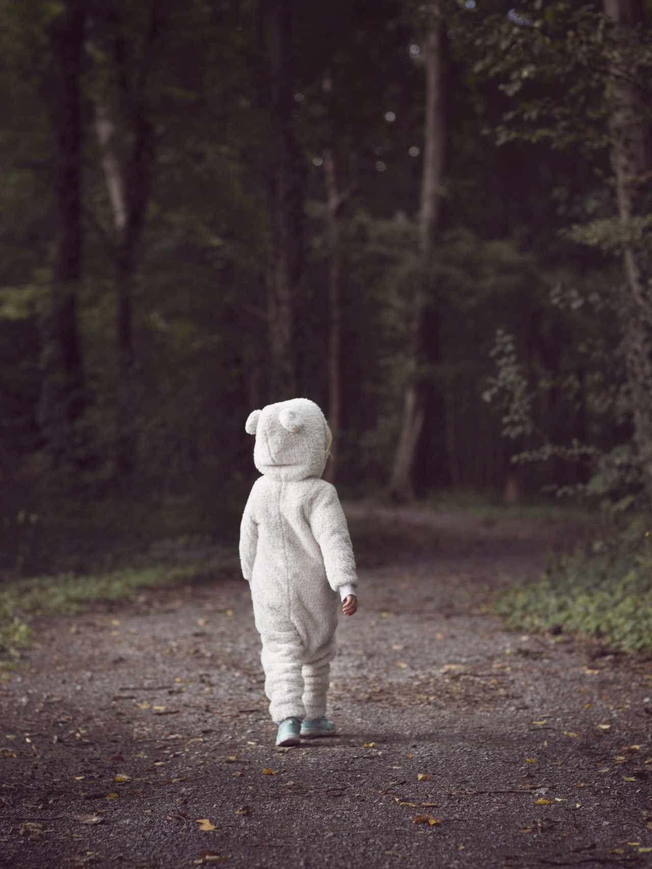 Small child wearing white bear suit, walking along a tree-lined forest path