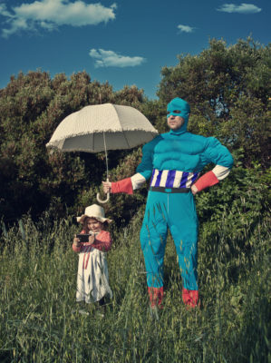 Man dressed as a super hero is protecting a little girl with umbrella.