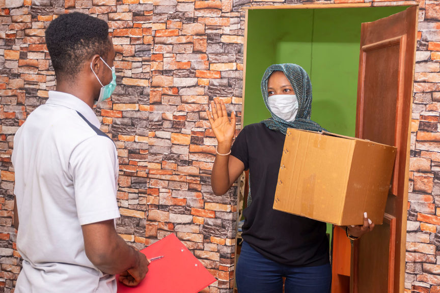 Woman receiving a parcel from a delivery service worker, wearing a face mask, waves the courier