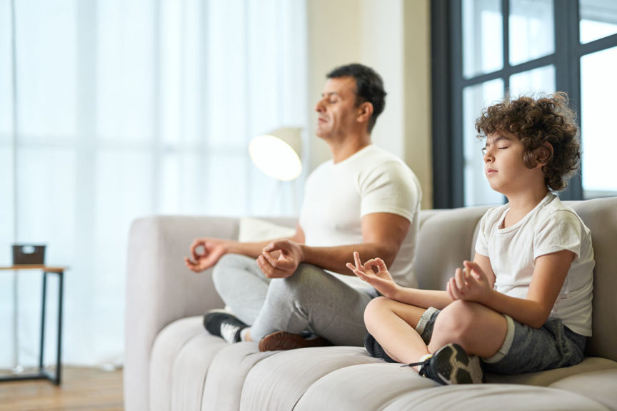 father and his son meditating together on a couch at home