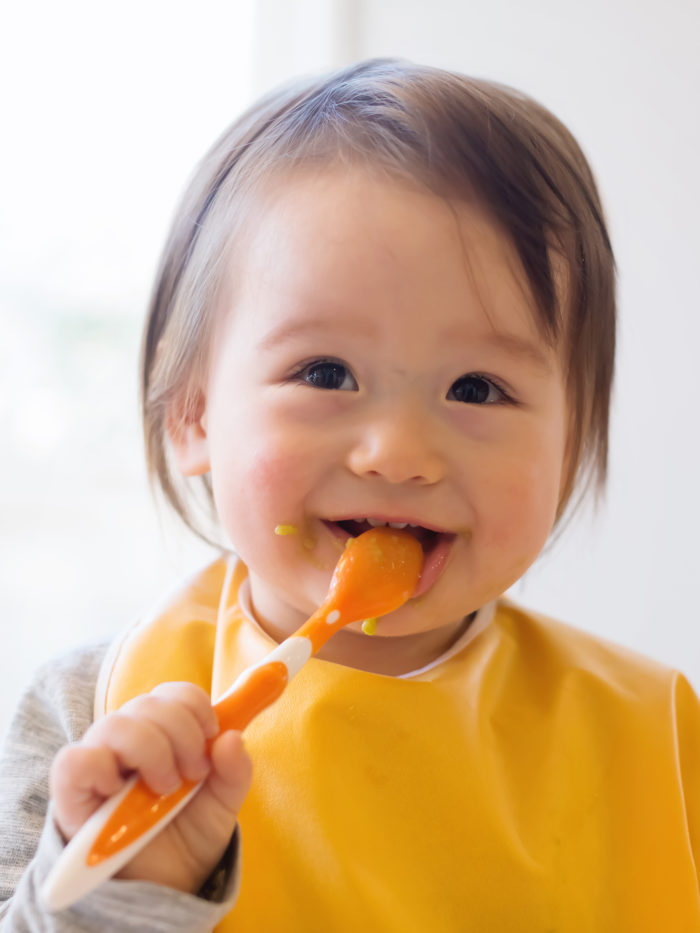 Baby with spoon smiling and enjoying baby food.