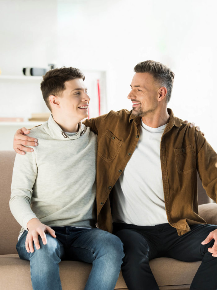 happy father and teen son hugging on sofa at home, looking at each other
