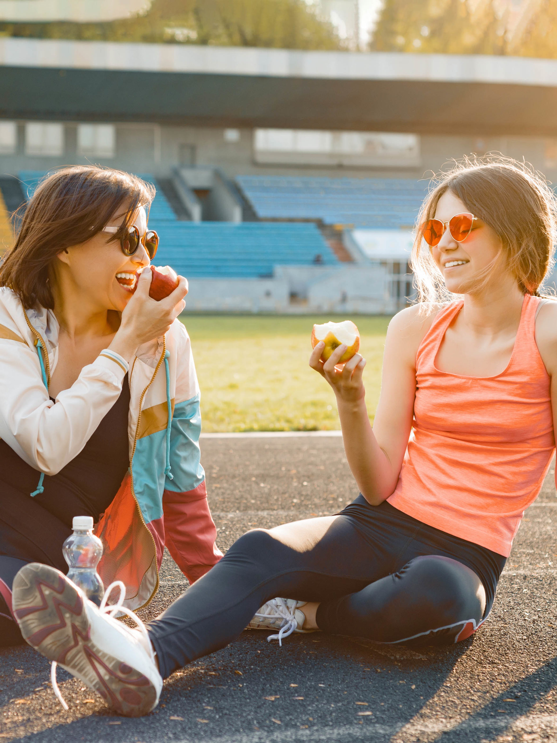 mother and teen daughter together eating apples on track field