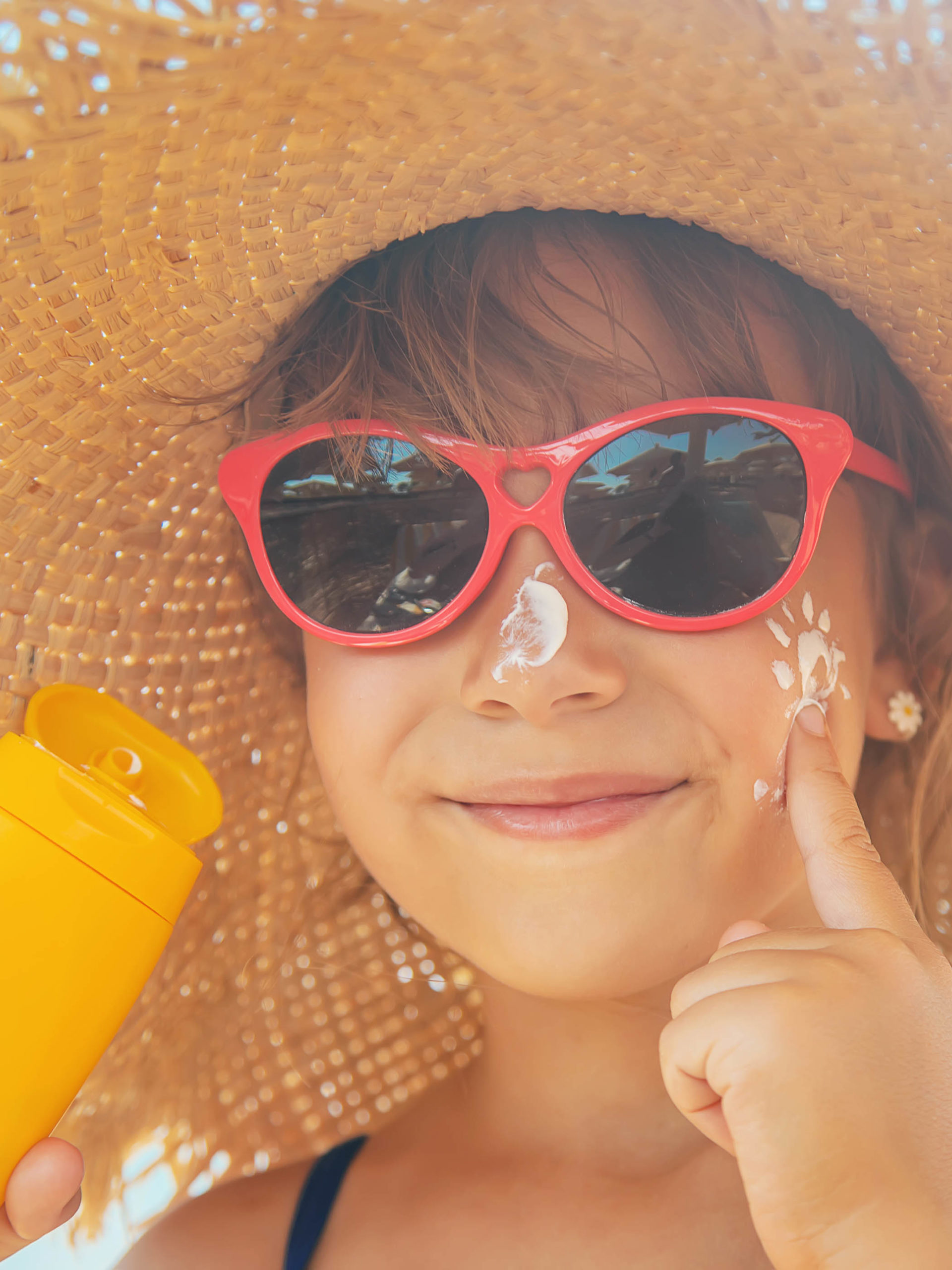 little girl with sunglasses and a hat applying sunscreen to her face