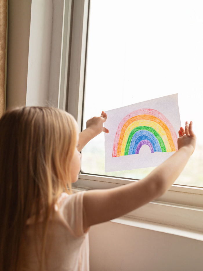 young girl hanging a picture of a rainbow she drew on a window