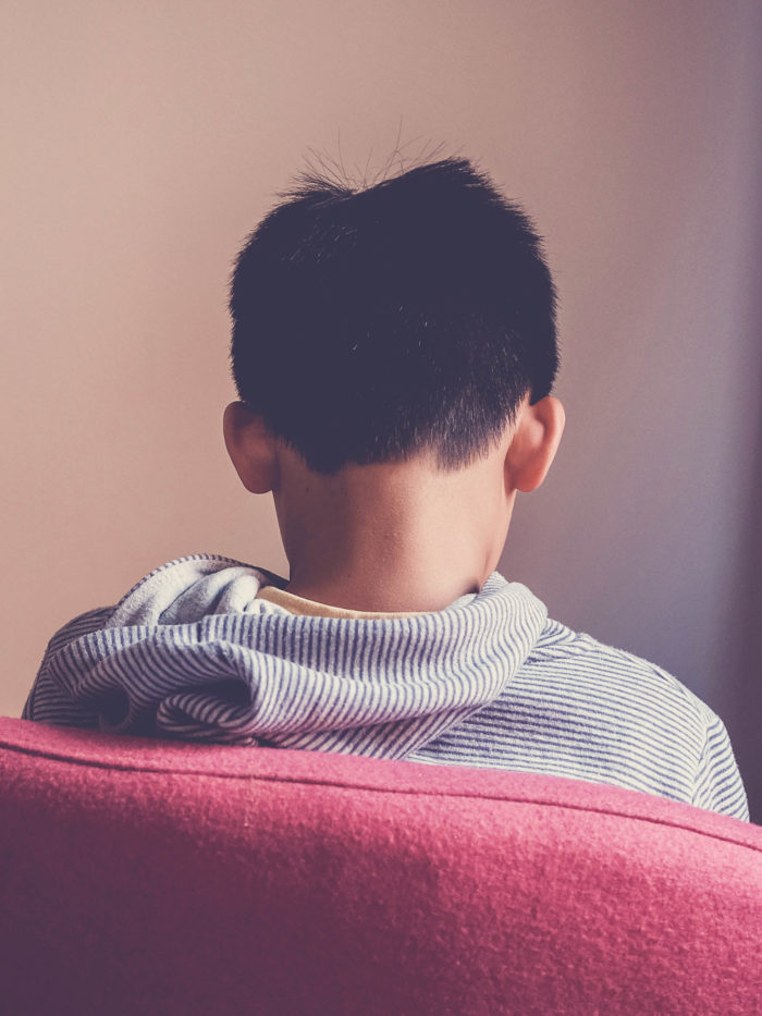 Sad preteen boy sitting alone in chair facing wall, depressed