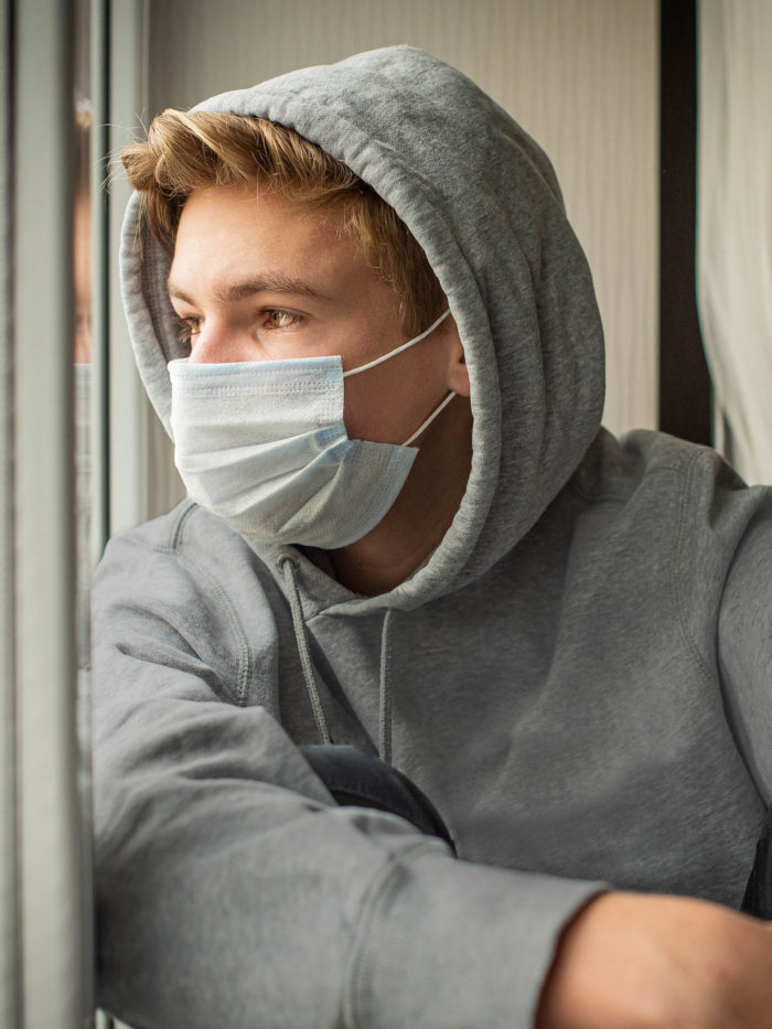 sad teenage boy wearing a mask during pandemic looking out the window