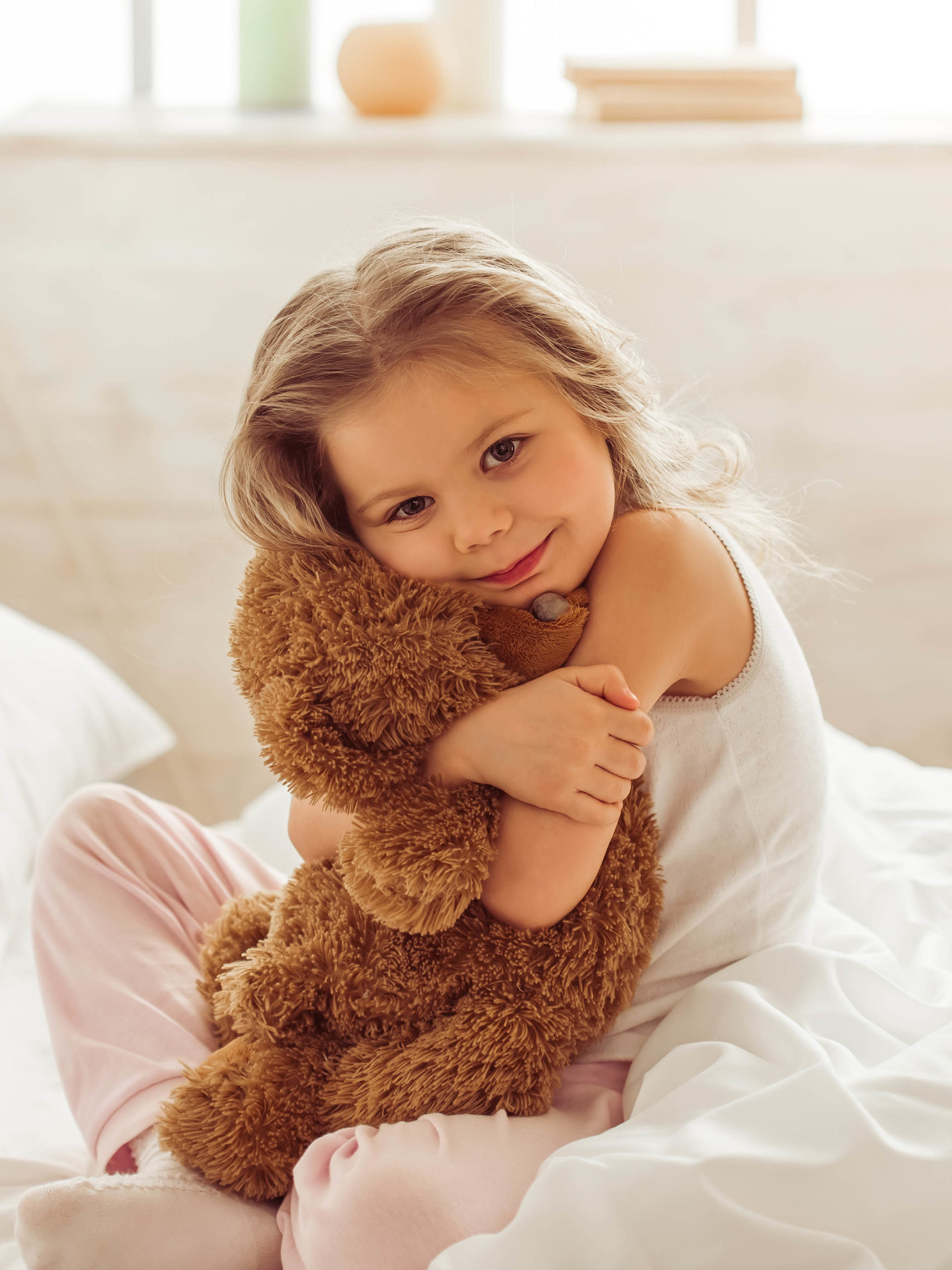Sweet little girl is hugging a teddy bear while sitting on her bed at home