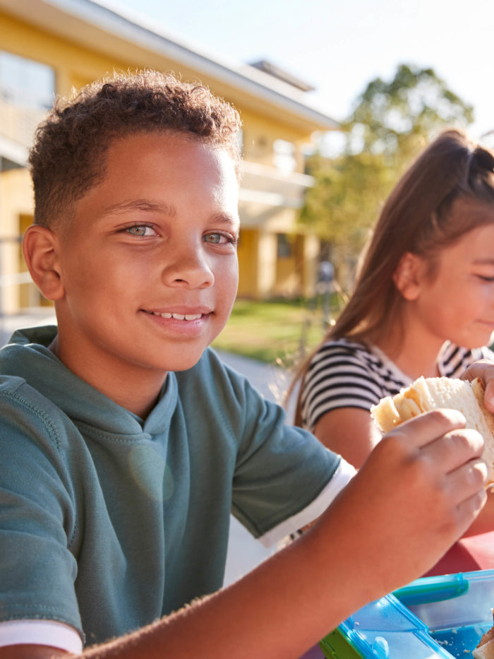 Boy at elementary school lunch table about to bite into sandwich for lunch