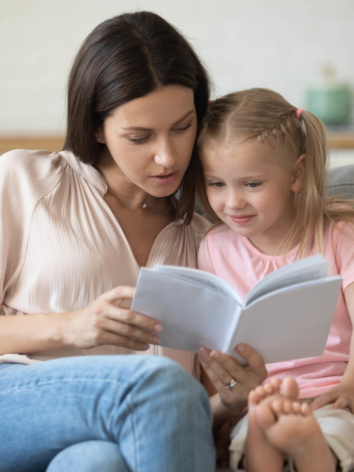 Young mother and preschool daughter sitting on couch, mom holding book reading