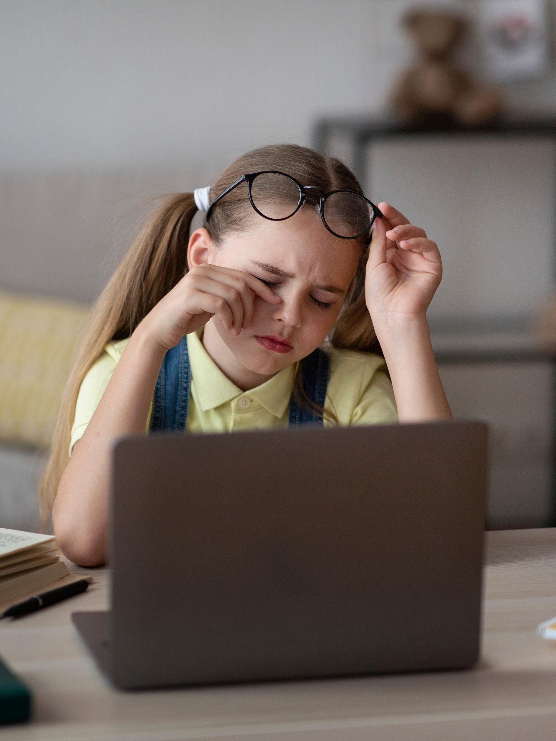 cranky girl taking off glasses, rubbing her eyes, sitting at table working on laptop
