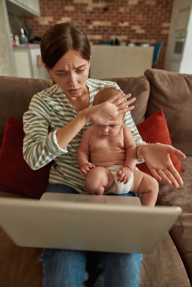 young worried mom with laptop holding infant while feeling his temperature with her hand on his forehead