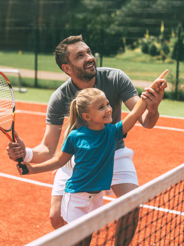 cheerful dad and daughter on tennis court, dad showing daughter how to play