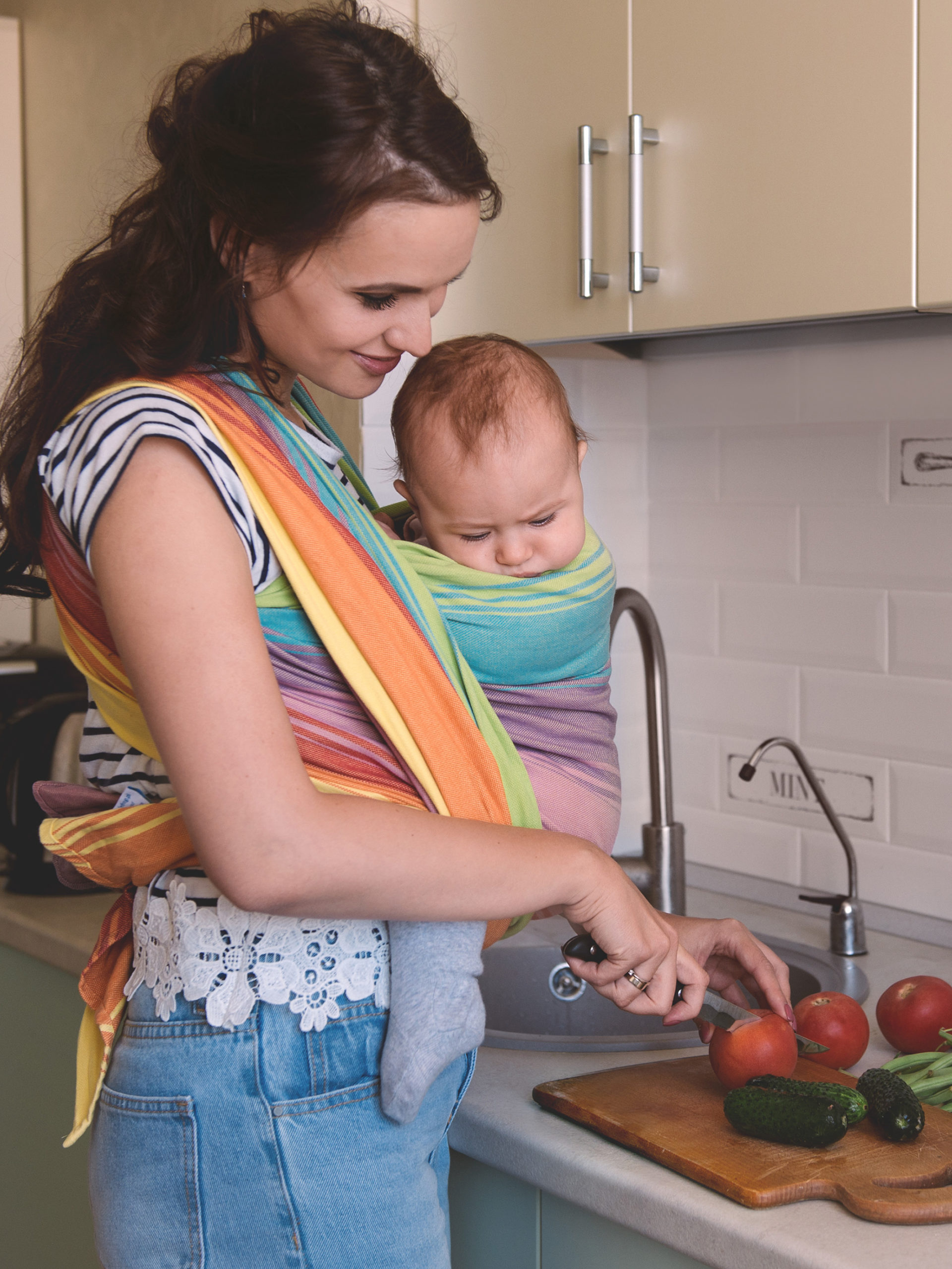 Young mom cooking in the kitchen with the little baby in a sling