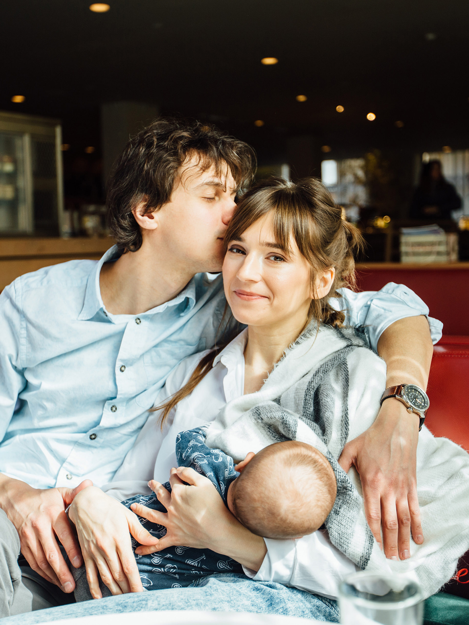 young happy family with husband kissing his wife while she is breast feeding their newborn little son in a restaurant