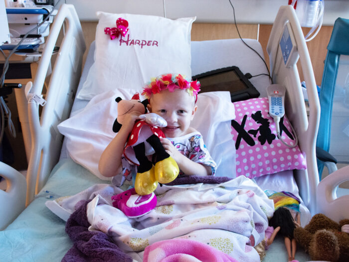 Harper sits in hospital bed and smiles while holding minnie mouse stuffed animal