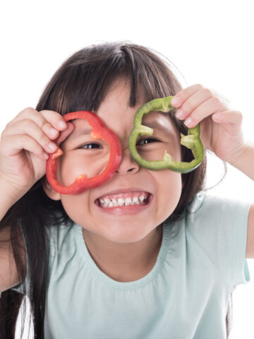 Child holds bell pepper rounds over eyes and smiles