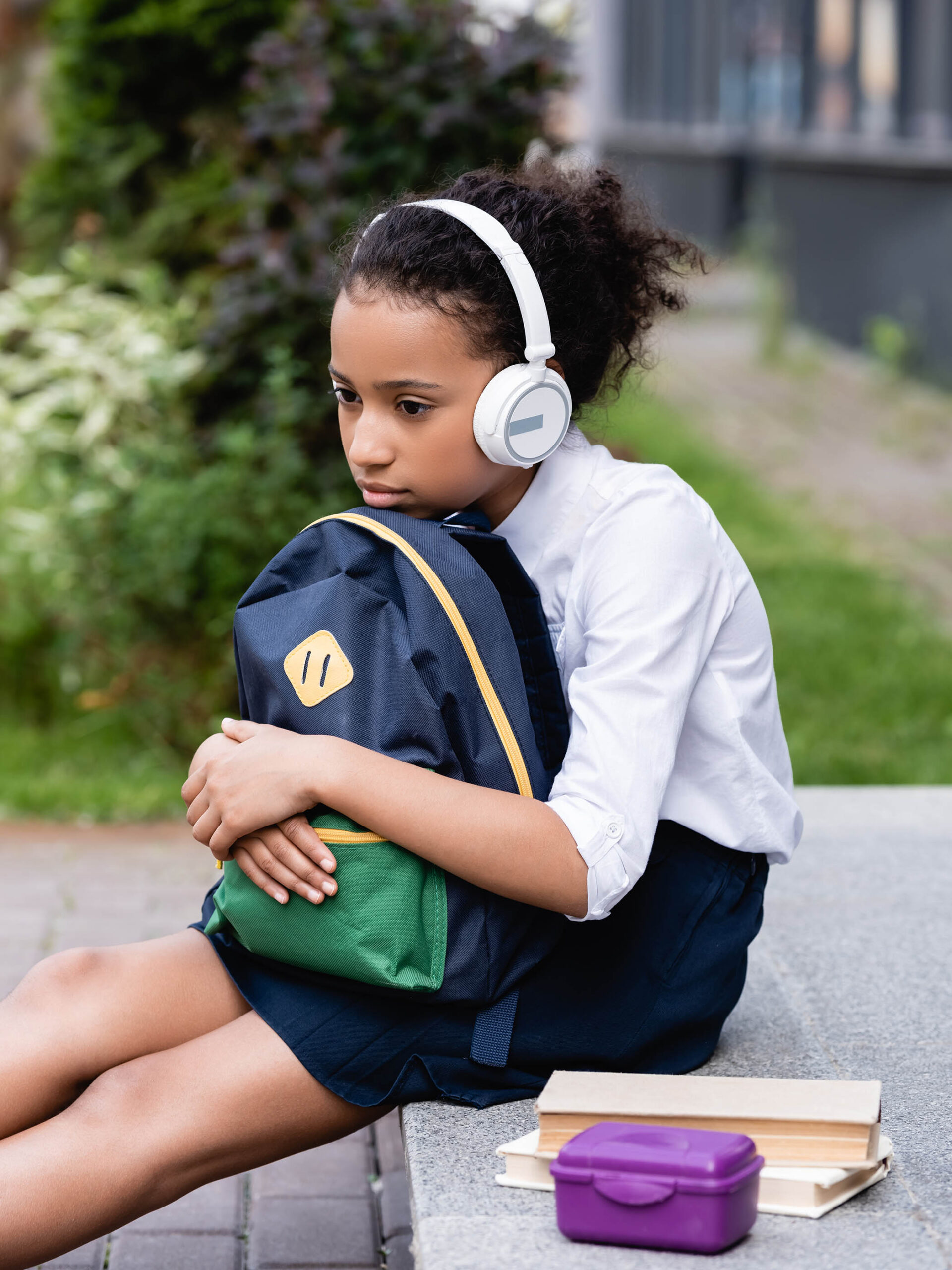 sad african american schoolgirl with backpack and headphones sitting outside