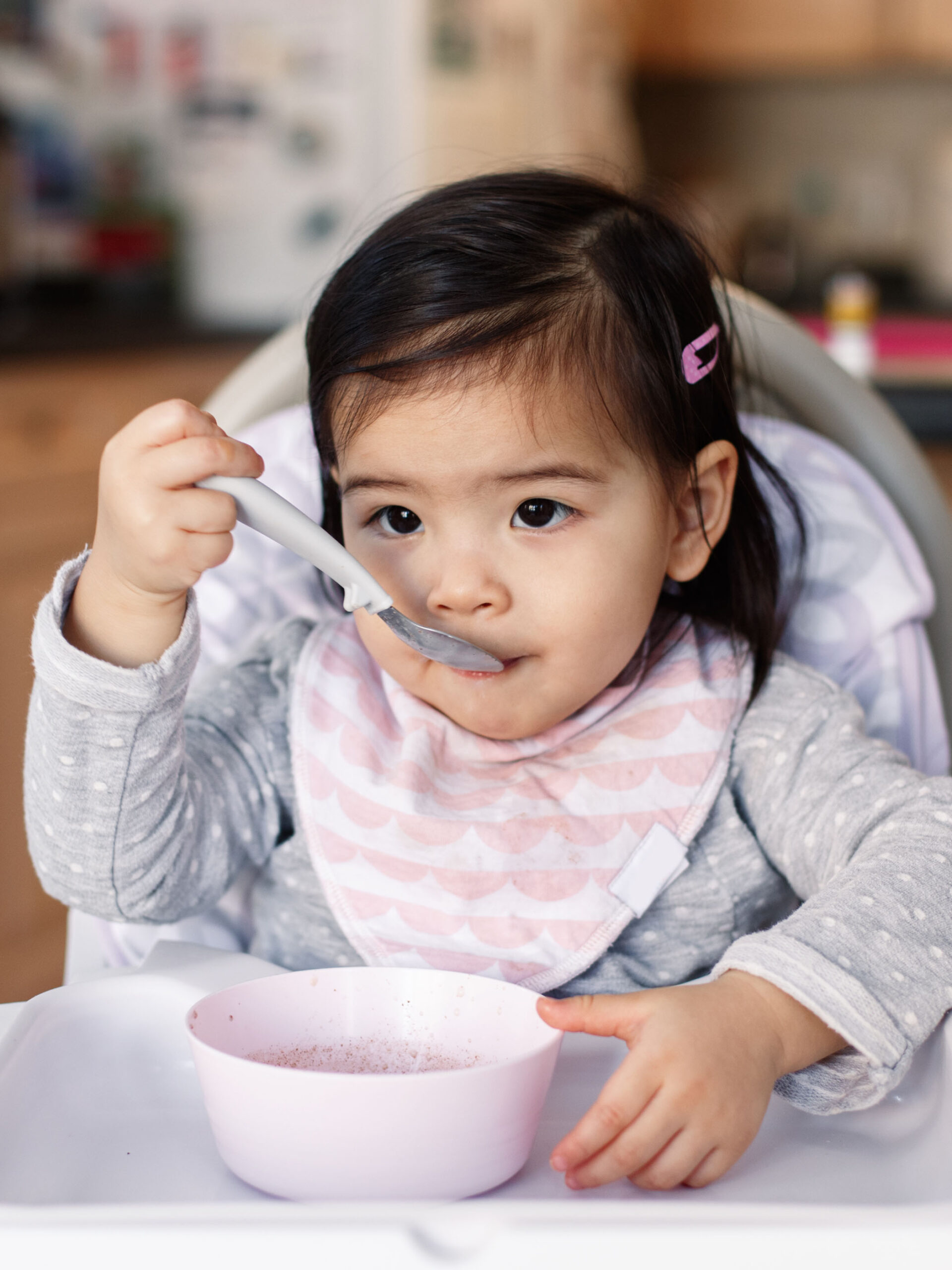 Cute adorable Asian Chinese kid girl sitting in high chair eating soup with spoon. Healthy eating for kids children. Toddler eating independently in kitchen at home. Candid real authentic moments.