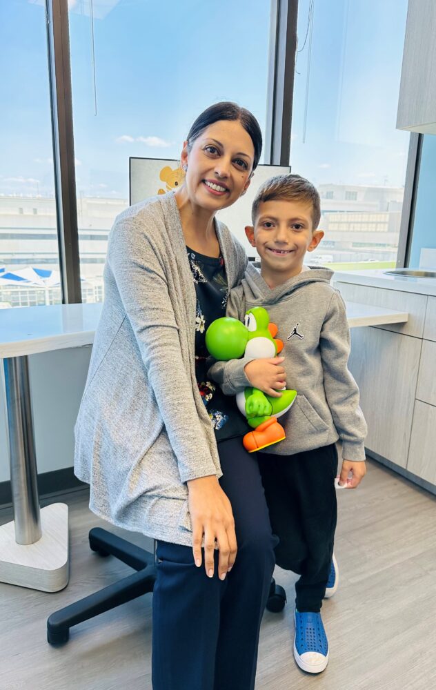 Ethan, wearing gray sweatshirt and holding Yoshi toy, stands next to Dr. Doshi and smiles 