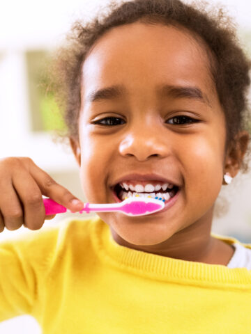 Young child smiles while brushing her teeth, look right into the camera, wearing yellow