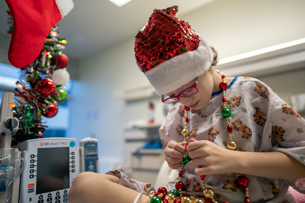 Patient celebrates holidays at CHOC hospital in orange - young girl in hospital gown wearing santa hat looks at ornament beads that she's wearing around her neck 