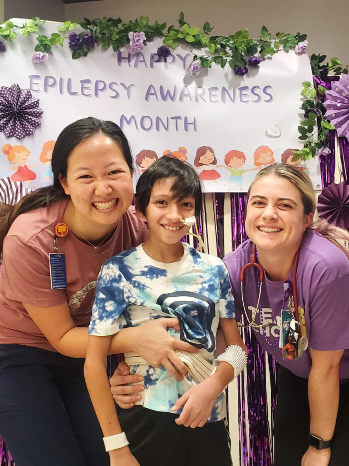 Iffers, Isa and Lauren smile together in front of Epilepsy Awareness Month banner 