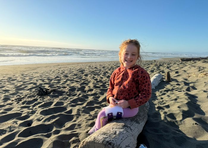 little girl with braces on legs sits on a log on the beach with the ocean and sky in the background