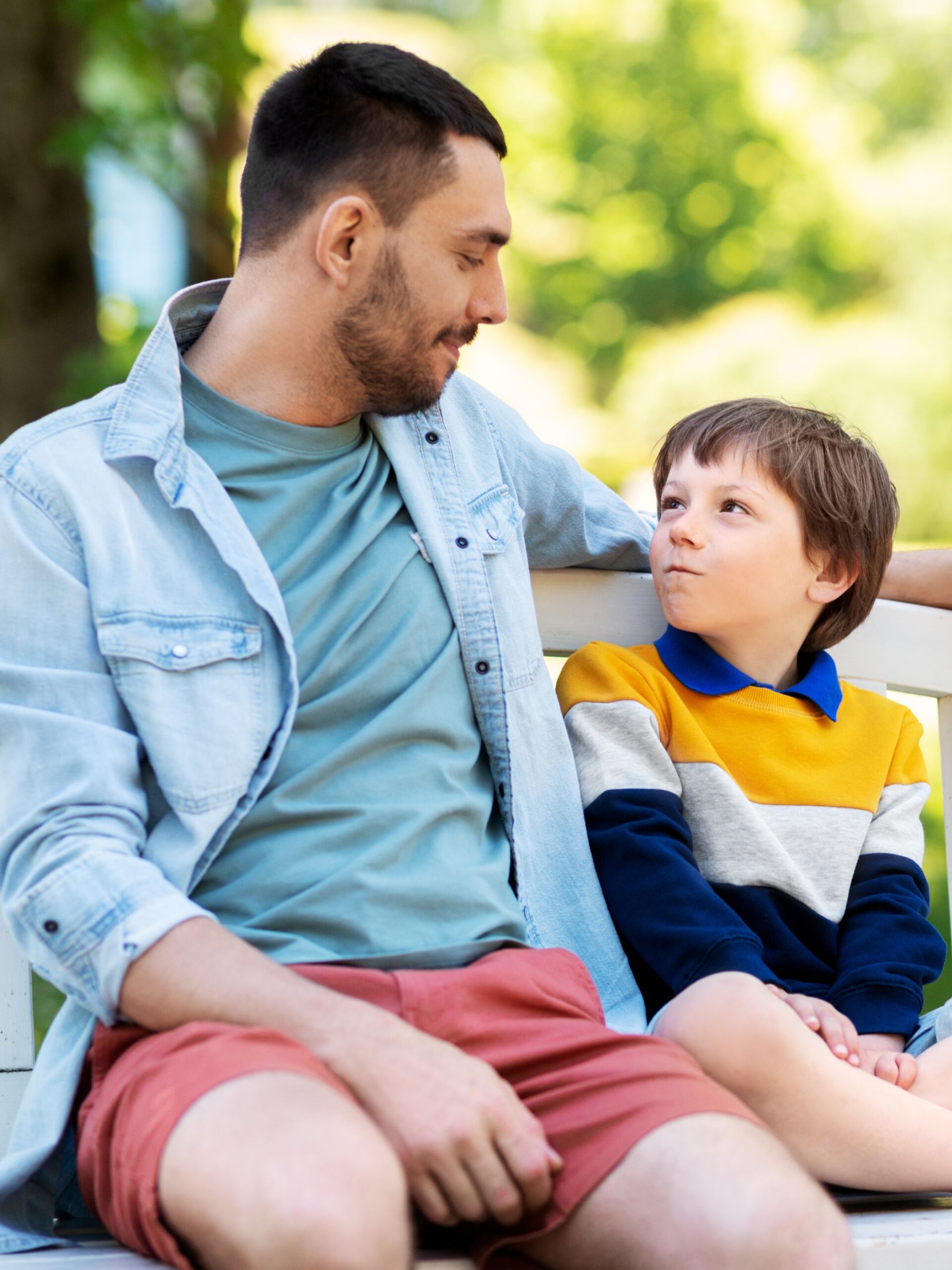 A man in a blue shirt and red shorts sits and talks with a boy with brown hair and a yellow and blue striped shirt