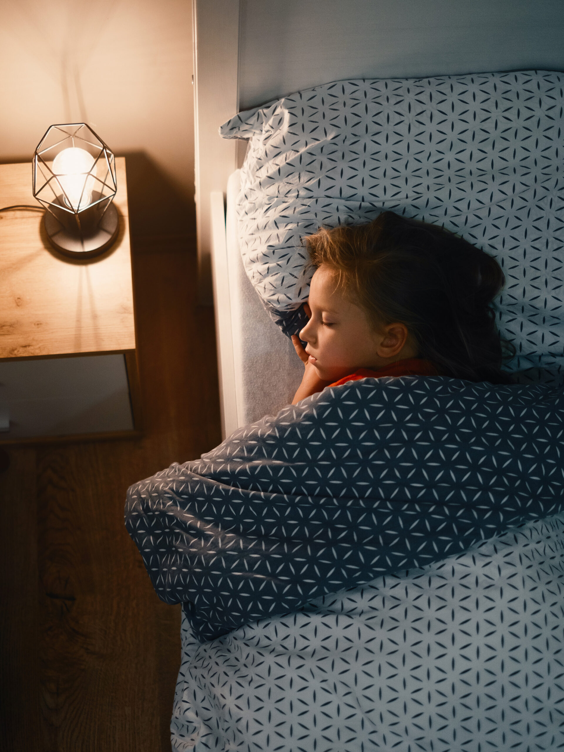 Child sleeps in bed with light on the nightstand