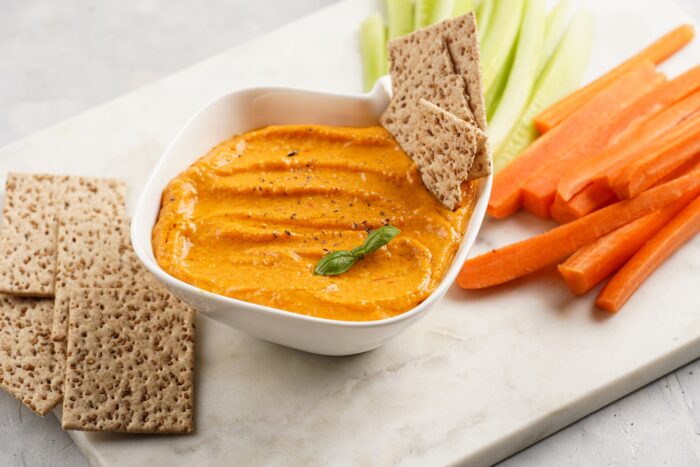 Paprika bell-pepper hummus dip, thin crackers with sesame seeds and carrot and cucumber cut in long strips and sticks