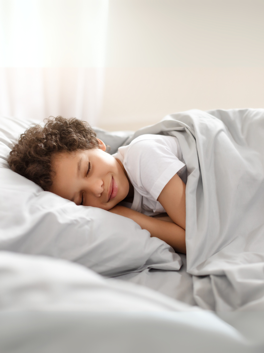 African American young boy peacefully sleeps in a bed, with his body curled up and his eyes closed. The room is dimly lit, and a blanket covers him.