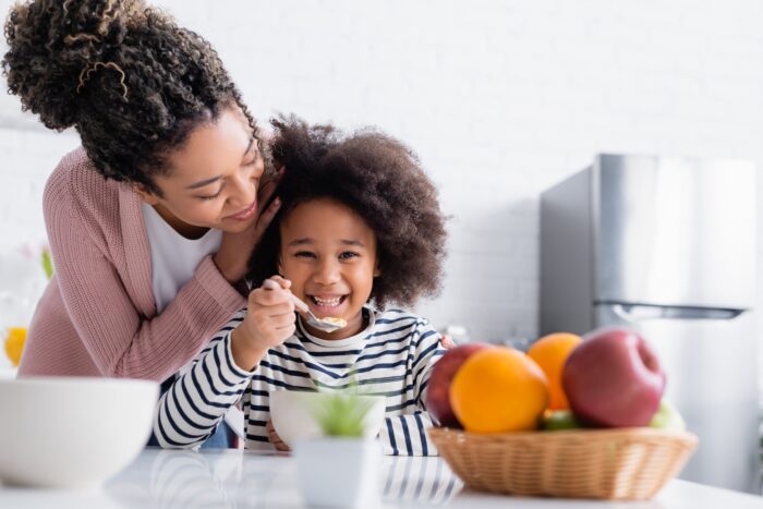african american woman touching hair of happy kid eating corn flakes near fruits on blurred foreground