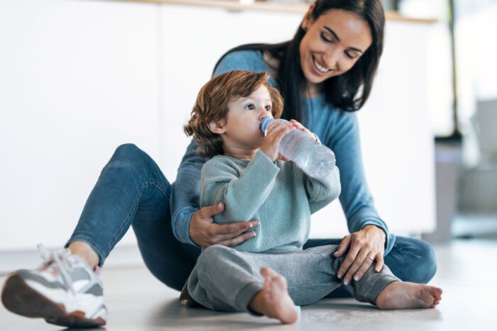 Shot of mother looking to her son while he drinking water from the plastic bottle sitting on the floor at home.