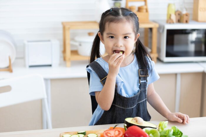 cute asian little child girl eating slice a cucumber with different vegetables in the kitchen