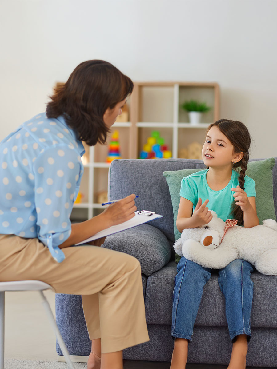Supportive psychologist with clipboard listening to little child during therapy session. Preschool girl feeling at ease in therapist's office sharing her thoughts and concerns