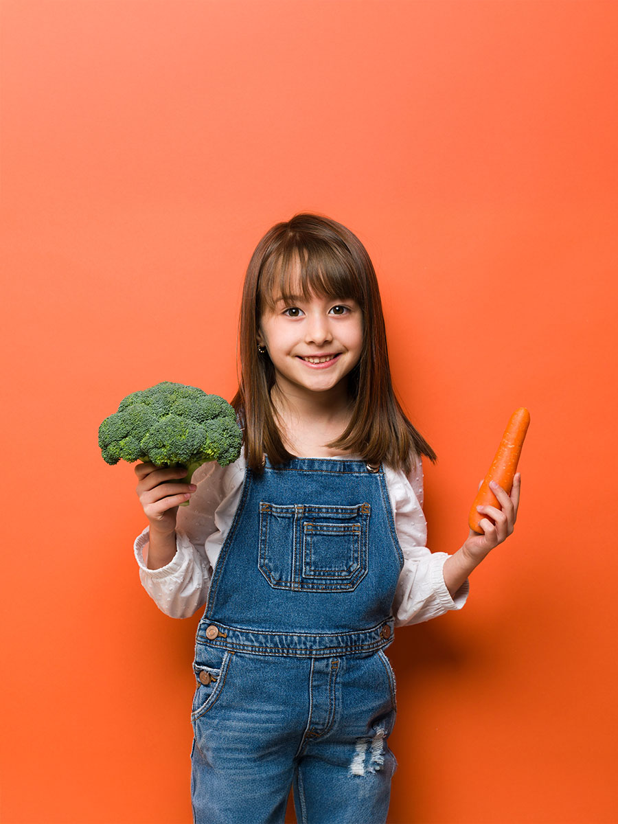 Cute looking little girl dressed casual and holding a carrot and some broccoli to show she eats a lot of healthy food