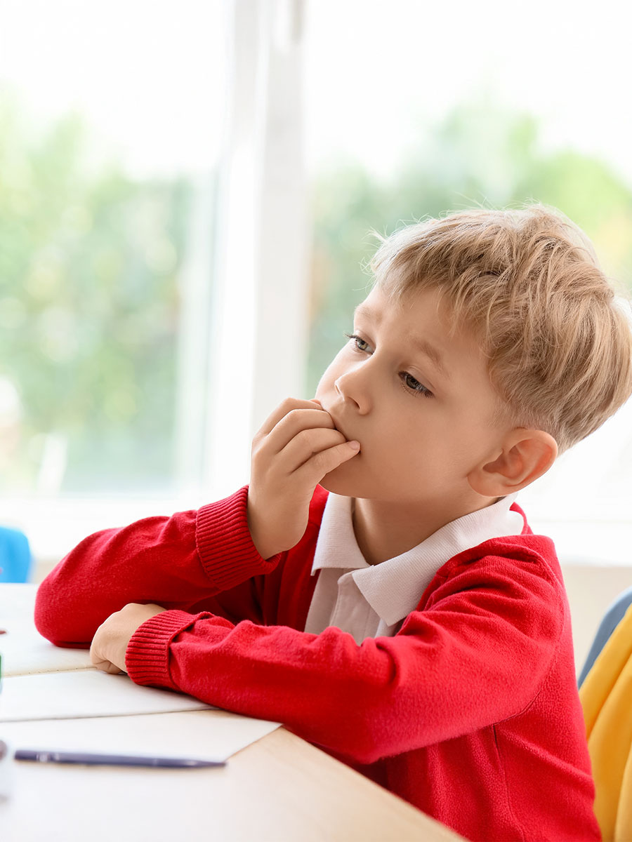 Little schoolboy biting nails at desk in classroom