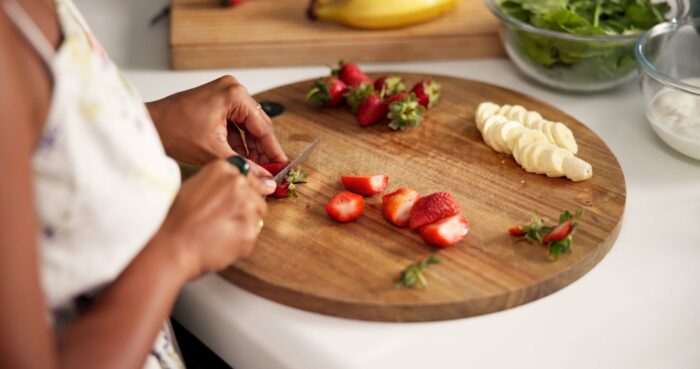 Woman, hands and cutting with fruit for prep, salad or healthy diet in kitchen at home. Meal closeup, female person or food preparation with natural organic strawberry or banana for vitamin nutrition
