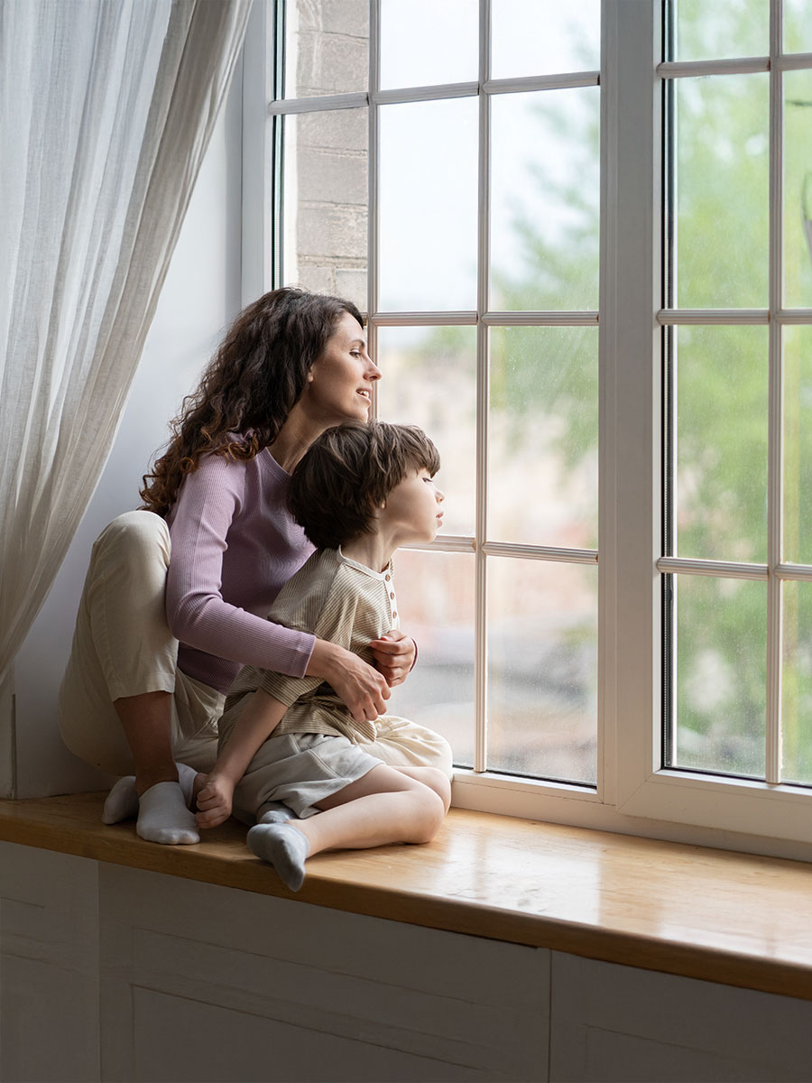 Curious mom and kid boy looking outside sitting on window sill. Caring mother or babysitter exploring world from inside on quarantine at home. Young mum and child son wait for dad return from work