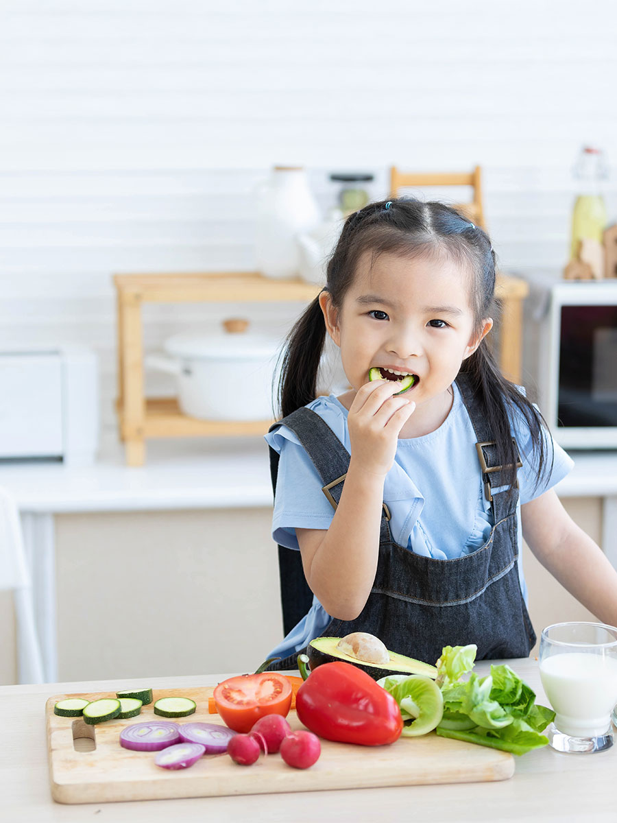 cute asian little child girl eating slice a cucumber with different vegetables in the kitchen