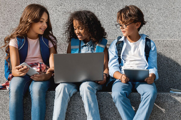 Smiling school kids preteen boys and girls elementary middle pupils children sitting on the stairs and looking in smartphone screen with digital devices laptop tablet in hands outdoors, copy space