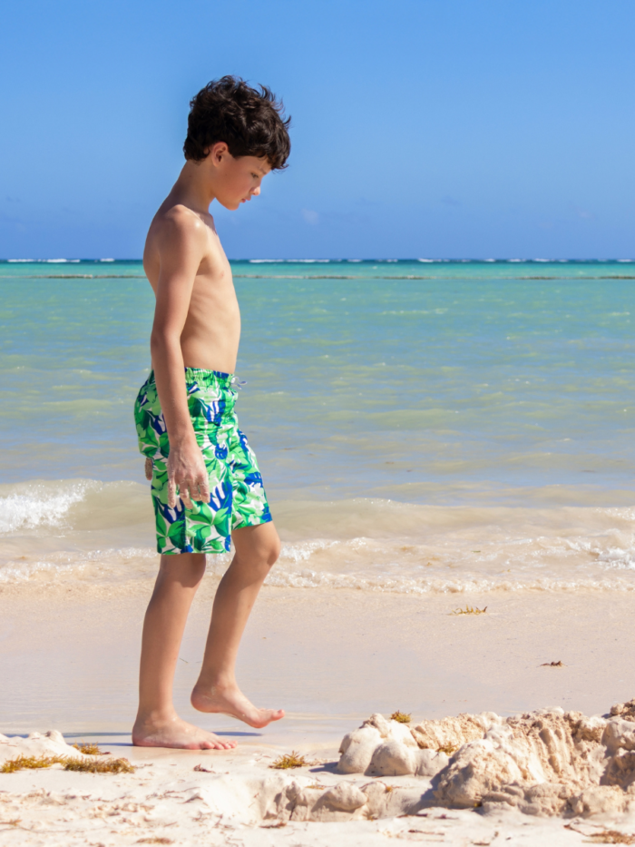 Teenager boy is playing with sand at the tropical beach, turquoise waters of the ocean are on the background, hot sunny day at the shore.