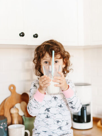 Little girl wearing pajamas drinking milk sitting on kitchen table in the morning. Foucs on hands holding glass of milk